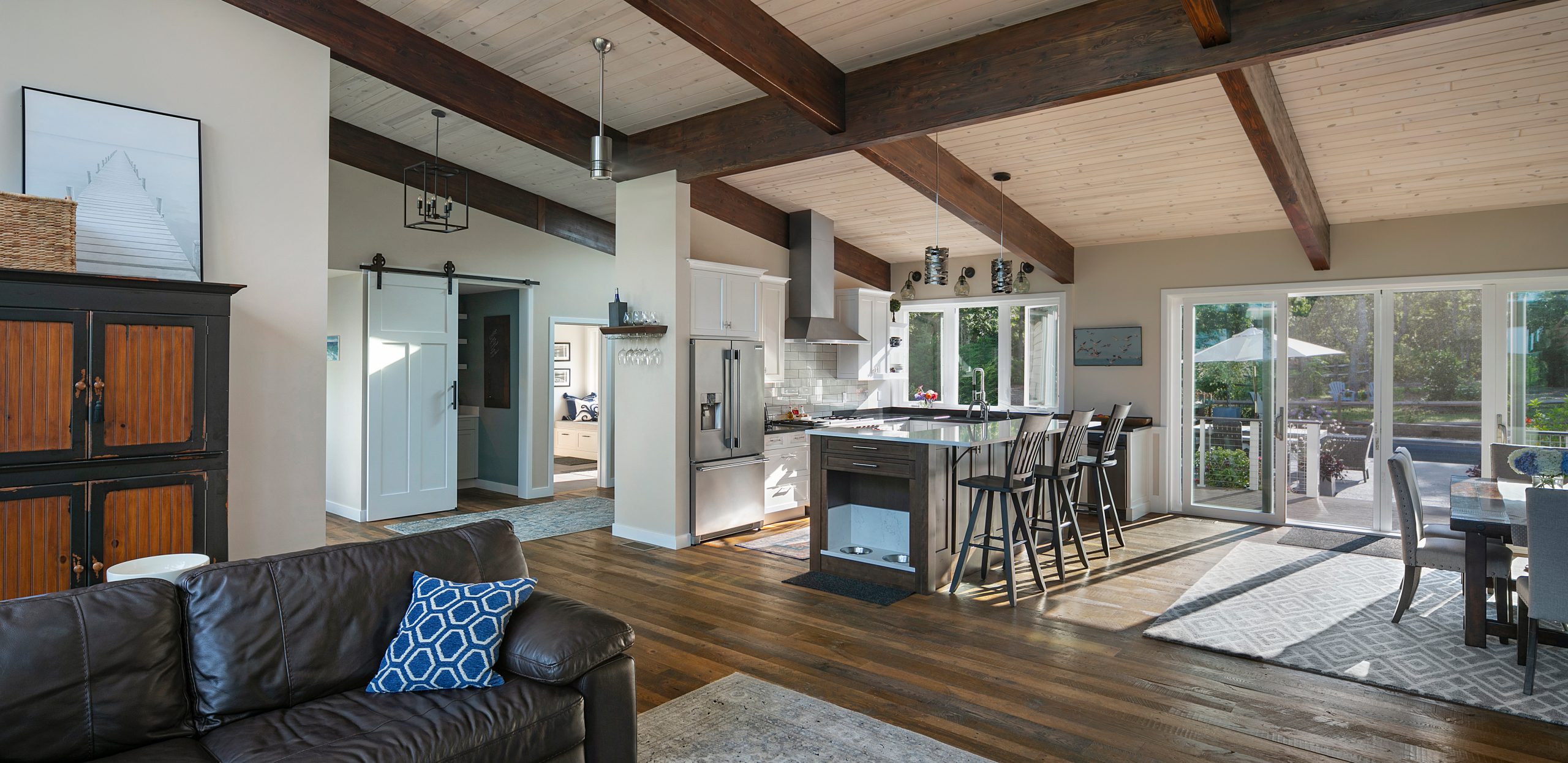 Truro Treehouse by Acorn Deck House Company is a modern farmhouse on Cape Cod. This photo shows a large open-concept living room, dining room, and kitchen with exposed post-and beam ceilings. The kitchen cabinets are white, the appliances are stainless steel, and the island is dark brown wood. The island features a cutout for dog bowls.